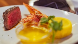A gourmet dish featuring a piece of seared steak cooked to medium rare on the left side of the plate, accompanied by a lobster tail in the middle. On the right, there is a small bowl of melted butter and a lemon wedge, garnished with a sprig of parsley.