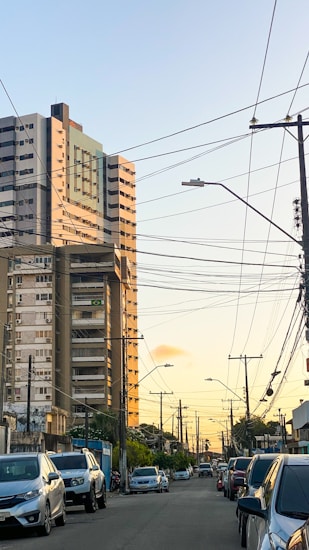 A city street during sunset with parked cars lining both sides. High-rise buildings are on the left, with the tallest building prominently featuring a Brazilian flag. Numerous power lines crisscross the sky, and streetlights are visible throughout the scene.