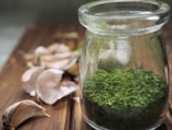 A glass jar containing dried green herbs is placed on a wooden surface, with several garlic cloves scattered around. The scene suggests a rustic kitchen setting.