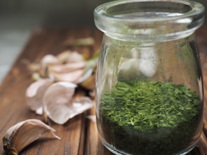 A glass jar containing dried green herbs is placed on a wooden surface, with several garlic cloves scattered around. The scene suggests a rustic kitchen setting.