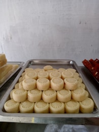 A metal tray is filled with neatly arranged pieces of round, creamy-colored sweets, each with a circular pattern on top. These are placed on a grey surface with a plain, textured background, and more trays with similar and other types of sweets can be seen partially to the sides.