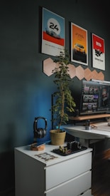 A modern workspace featuring a white drawer unit with a wooden top, set against a dark wall. The top holds a pair of headphones resting on a wooden stand, a potted plant with leafy foliage, a black camera placed on a small tray, and a partially completed jigsaw puzzle. In the background, a desk with a large computer monitor displaying various images and videos flanks the wall. Above the desk, posters related to car racing, including imagery and text such as '24 Heures du Mans' and 'James Hunt', are displayed. Geometric wooden wall decorations add to the aesthetic.