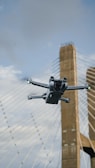 A drone is flying in the foreground with a suspension bridge in the background. The bridge towers are visible, and the scene is set against a partly cloudy sky.