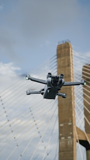 A drone is flying in the foreground with a suspension bridge in the background. The bridge towers are visible, and the scene is set against a partly cloudy sky.
