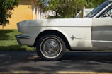 A side view of the front half of a vintage white Mustang car is parked on the side of a road. The chrome details and iconic emblem are prominently featured, with a grassy area and part of a yellow building visible in the background.