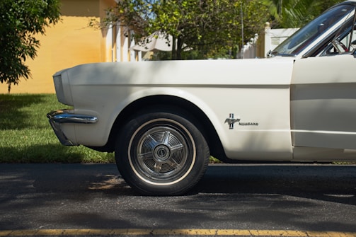 A side view of the front half of a vintage white Mustang car is parked on the side of a road. The chrome details and iconic emblem are prominently featured, with a grassy area and part of a yellow building visible in the background.