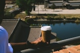 A person holds a takeaway coffee cup while leaning on a railing, overlooking a tranquil setting with a body of water and traditional-style buildings in the background. The scene is calm and bathed in natural sunlight, casting shadows and creating reflections on the water's surface.