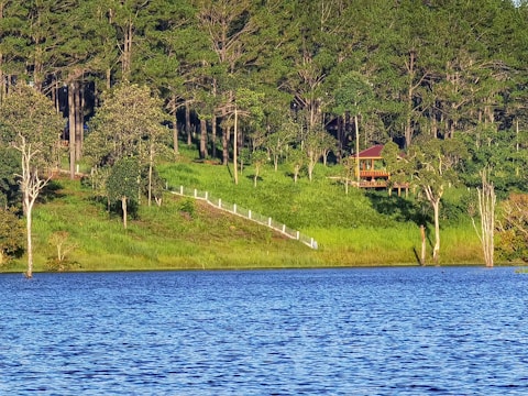 A serene lakeside view with a lush green forest in the background. A wooden cabin with a red roof is nestled among the trees. A path with white railings descends from the forest to the lake. The water reflects the rich blue of the sky.
