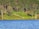 A serene lakeside view with a lush green forest in the background. A wooden cabin with a red roof is nestled among the trees. A path with white railings descends from the forest to the lake. The water reflects the rich blue of the sky.