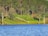 A serene lakeside view with a lush green forest in the background. A wooden cabin with a red roof is nestled among the trees. A path with white railings descends from the forest to the lake. The water reflects the rich blue of the sky.