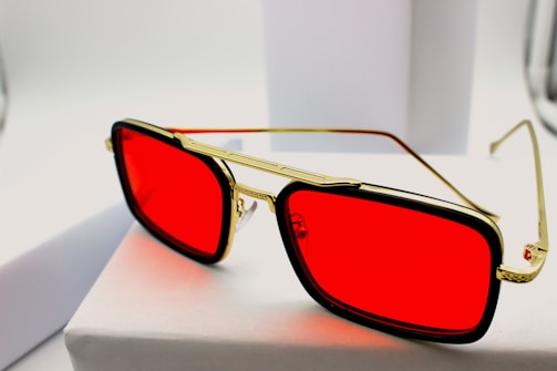 A pair of stylish sunglasses with bold red lenses rests on a white surface. The frame is a combination of gold and black, giving the eyewear a luxurious and modern appearance.