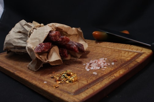 A rustic setup on a wooden cutting board with a bundle of sausages wrapped in brown paper, pink Himalayan salt, and a mix of herbs and spices. A knife with a black handle and a knife blade is visible, along with a half-cut tomato in the background.