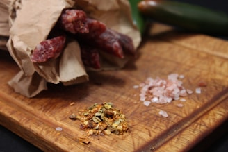 A rustic wooden cutting board holds a partially unwrapped bundle of cured sausages in brown paper alongside piles of pink Himalayan salt and mixed spices. A jalapeño pepper is placed in the background, adding a fresh green contrast.