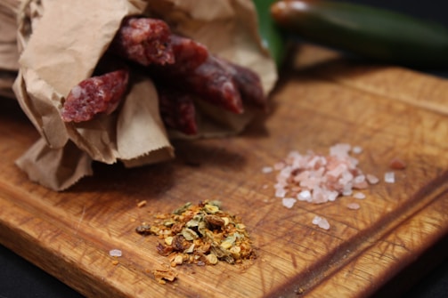 A rustic wooden cutting board holds a partially unwrapped bundle of cured sausages in brown paper alongside piles of pink Himalayan salt and mixed spices. A jalapeño pepper is placed in the background, adding a fresh green contrast.