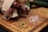 A rustic wooden cutting board holds a partially unwrapped bundle of cured sausages in brown paper alongside piles of pink Himalayan salt and mixed spices. A jalapeño pepper is placed in the background, adding a fresh green contrast.