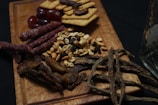 A wooden serving board holds an assortment of snacks including red grapes, crackers, thin dried sausage sticks, peanuts, raisins, and thin strips of dried meat. The background is dark, enhancing the contrast of the food items.