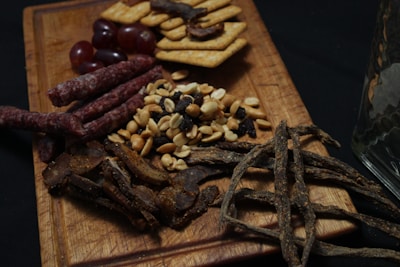 A wooden serving board holds an assortment of snacks including red grapes, crackers, thin dried sausage sticks, peanuts, raisins, and thin strips of dried meat. The background is dark, enhancing the contrast of the food items.