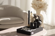 A stylish modern setting featuring a light-colored armchair and a minimalist arrangement on a pinkish table. The table displays a chic decorative setup with black and white sculptural candles and a black vase filled with dried flowers atop a large book titled 'TOM FORD'. Soft natural light filters through sheer curtains, casting gentle shadows.