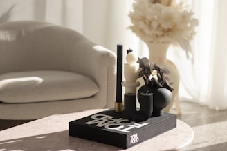 A stylish modern setting featuring a light-colored armchair and a minimalist arrangement on a pinkish table. The table displays a chic decorative setup with black and white sculptural candles and a black vase filled with dried flowers atop a large book titled 'TOM FORD'. Soft natural light filters through sheer curtains, casting gentle shadows.