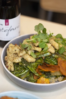 A bowl filled with a vibrant and colorful dish containing various vegetables such as broccoli, garnished with green herbs. There are also pieces of cooked chicken seasoned with pepper on top. The dish is placed on a light-colored surface, and beside it is a bottle of wine.
