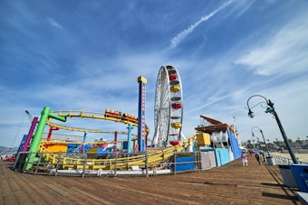 A vibrant amusement park features a large Ferris wheel with red and yellow cabins, a colorful roller coaster with green, yellow, and red tracks, and various structures set against a clear blue sky. Street lamps and people are visible along a wooden boardwalk, adding to the lively seaside atmosphere.