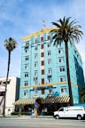 A vibrant, multi-story hotel with an art deco style, painted in a striking turquoise color. The top portion is accented with yellow and features decorative designs around the windows. There's a U.S. flag on the roof. Two tall palm trees are in front, one on each side of the building. A white van is parked in front of the hotel near the street, and the building has an entrance with a striped awning.