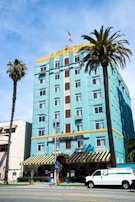 A vibrant, multi-story hotel with an art deco style, painted in a striking turquoise color. The top portion is accented with yellow and features decorative designs around the windows. There's a U.S. flag on the roof. Two tall palm trees are in front, one on each side of the building. A white van is parked in front of the hotel near the street, and the building has an entrance with a striped awning.
