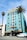 A vibrant, multi-story hotel with an art deco style, painted in a striking turquoise color. The top portion is accented with yellow and features decorative designs around the windows. There's a U.S. flag on the roof. Two tall palm trees are in front, one on each side of the building. A white van is parked in front of the hotel near the street, and the building has an entrance with a striped awning.