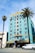 A vibrant, multi-story hotel with an art deco style, painted in a striking turquoise color. The top portion is accented with yellow and features decorative designs around the windows. There's a U.S. flag on the roof. Two tall palm trees are in front, one on each side of the building. A white van is parked in front of the hotel near the street, and the building has an entrance with a striped awning.