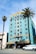 A vibrant, multi-story hotel with an art deco style, painted in a striking turquoise color. The top portion is accented with yellow and features decorative designs around the windows. There's a U.S. flag on the roof. Two tall palm trees are in front, one on each side of the building. A white van is parked in front of the hotel near the street, and the building has an entrance with a striped awning.