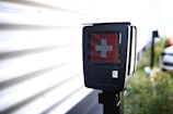 A black electronic device displaying a red screen with a white cross symbol in the center. It is mounted on a pole and located outdoors near a corrugated metal wall, with a blurred background that includes greenery and part of a vehicle.