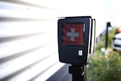 A black electronic device displaying a red screen with a white cross symbol in the center. It is mounted on a pole and located outdoors near a corrugated metal wall, with a blurred background that includes greenery and part of a vehicle.