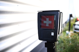 A black electronic device displaying a red screen with a white cross symbol in the center. It is mounted on a pole and located outdoors near a corrugated metal wall, with a blurred background that includes greenery and part of a vehicle.