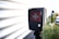 A black electronic device displaying a red screen with a white cross symbol in the center. It is mounted on a pole and located outdoors near a corrugated metal wall, with a blurred background that includes greenery and part of a vehicle.