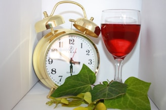 A gold-colored alarm clock with black numerals and hands sits next to a glass filled with red liquid, possibly wine. In front of the clock and glass are green leaves, adding a touch of nature to the scene.