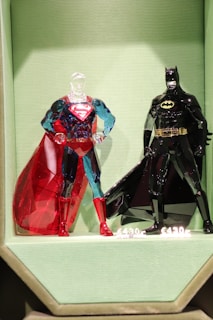 Two intricately designed glass figurines resembling well-known superheroes are displayed on a textured, light green background. The figure on the left is predominantly blue and red, featuring a transparent head and a vivid red cape. The figure on the right is almost entirely black with a yellow belt and logo, and a glossy black cape. Both figures are positioned upright with hands on hips.