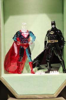 Two intricately designed glass figurines resembling well-known superheroes are displayed on a textured, light green background. The figure on the left is predominantly blue and red, featuring a transparent head and a vivid red cape. The figure on the right is almost entirely black with a yellow belt and logo, and a glossy black cape. Both figures are positioned upright with hands on hips.