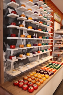 A display showcasing a variety of colorful cheeses on wall-mounted shelves and a wooden counter. The cheeses are circular and individually wrapped in vibrant colors like red, yellow, orange, purple, and green. Some display signs are present, and small serving bowls with spoons accompany the cheese samples. The background features a grayscale mural or image.