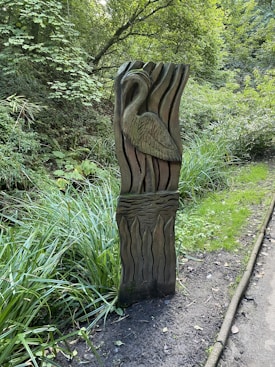 A wooden sculpture is situated in a lush, green forest area, depicting a bird in intricate detail against a backdrop of abstract wave patterns. The sculpture stands on a muddy path lined with overgrown grass and dense vegetation, with trees and bushes in the background creating a serene woodland atmosphere.