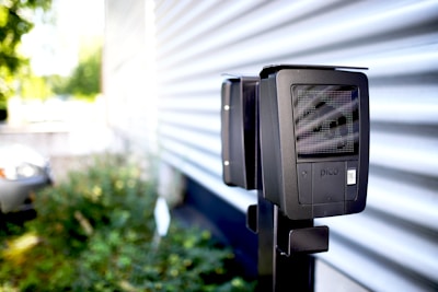 A black device is mounted on a metal pole in an outdoor setting with a corrugated metal wall in the background. The device has a screen and a label with the word 'pico'. There is greenery out of focus to the left, and part of a vehicle is barely visible in the background.
