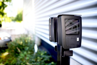 A black device is mounted on a metal pole in an outdoor setting with a corrugated metal wall in the background. The device has a screen and a label with the word 'pico'. There is greenery out of focus to the left, and part of a vehicle is barely visible in the background.