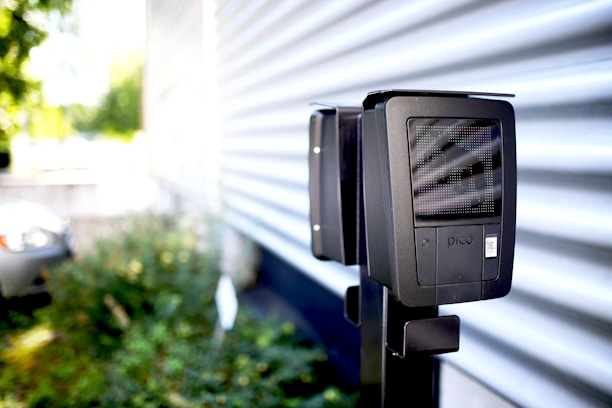 A black device is mounted on a metal pole in an outdoor setting with a corrugated metal wall in the background. The device has a screen and a label with the word 'pico'. There is greenery out of focus to the left, and part of a vehicle is barely visible in the background.