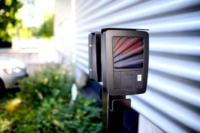 A digital parking meter or electric vehicle charging station is mounted on a post in an outdoor setting. It is located near a wavy, metallic wall. In the background, a car is partially visible along with some greenery, indicating an outdoor parking or urban environment.