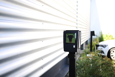 A black electronic device mounted on a pole is situated near a corrugated metal wall. The surroundings are outdoors, with plants and a white car in the background. There is bright natural lighting creating shadow patterns on the wall.