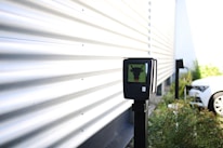 A black electronic device mounted on a pole is situated near a corrugated metal wall. The surroundings are outdoors, with plants and a white car in the background. There is bright natural lighting creating shadow patterns on the wall.