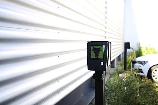 A black electronic device mounted on a pole is situated near a corrugated metal wall. The surroundings are outdoors, with plants and a white car in the background. There is bright natural lighting creating shadow patterns on the wall.