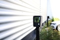 A black electronic device mounted on a pole is situated near a corrugated metal wall. The surroundings are outdoors, with plants and a white car in the background. There is bright natural lighting creating shadow patterns on the wall.