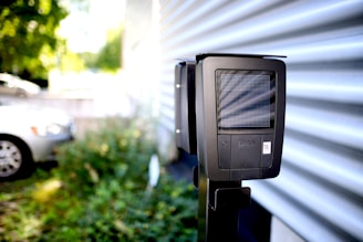 A dark-colored charging station with a digital display is installed outdoors next to a corrugated metal wall. In the background, a white car is partially visible in a parking area surrounded by greenery.