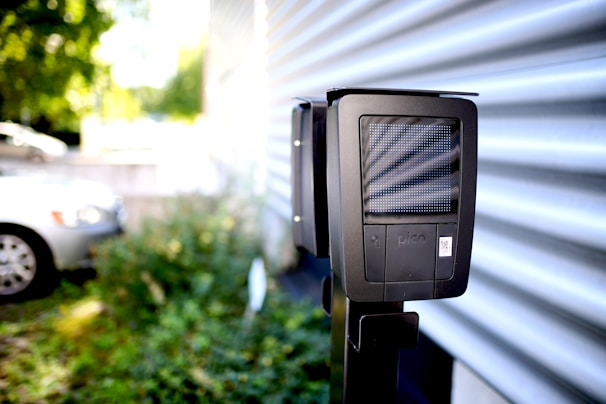 A dark-colored charging station with a digital display is installed outdoors next to a corrugated metal wall. In the background, a white car is partially visible in a parking area surrounded by greenery.