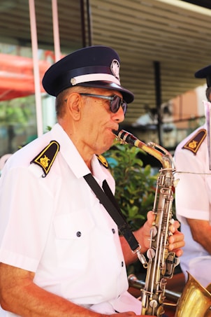 An older man is playing a saxophone while wearing a white uniform with dark shoulder patches and a dark blue cap with a badge. He is also wearing sunglasses. In the background, there are green plants and a blurred structure.